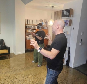 Tess standing in an open room with the AVP on their face and the battery pack in their pocket. Oliver is showing Tess the gestures for using the AVP.